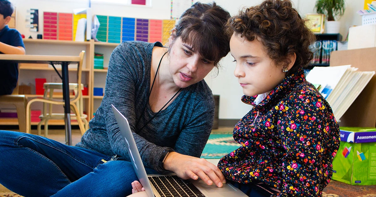 Montessori for All student works on computer with teacher. 