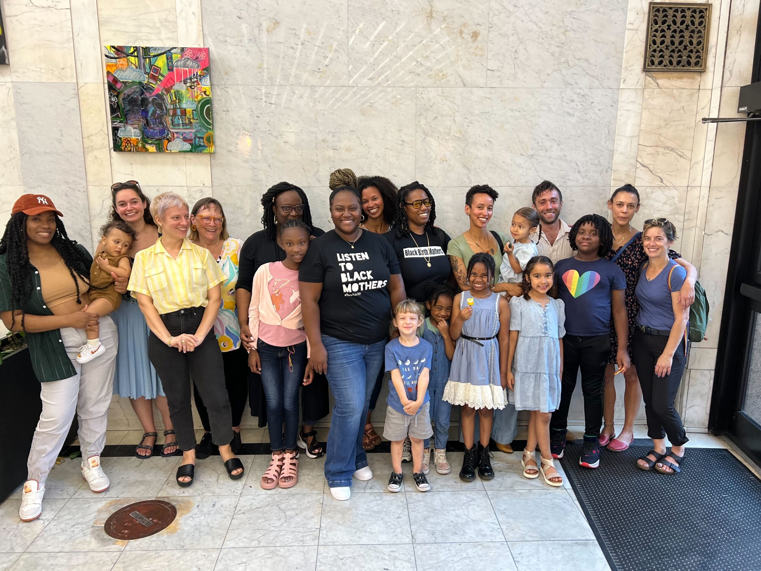 A group of women, men and children stand (with a couple of small children being held) outside of Birthmark Doula Collective, also known as New Orleans Breastfeeding Center. The worker-owned cooperative is focused on improving maternal health and perinatal outcomes for historically disinvested communities in New Orleans.