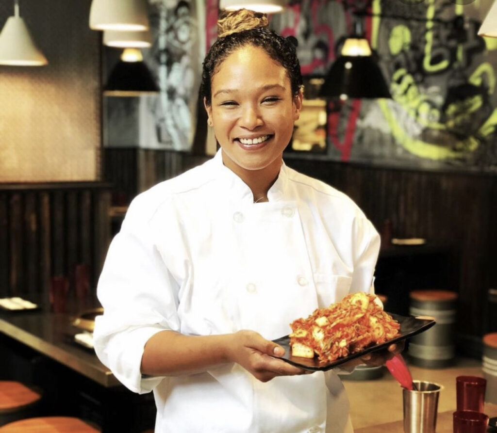 Female chef of color wearing white apron, holding a plate of food, and smiling at the camera