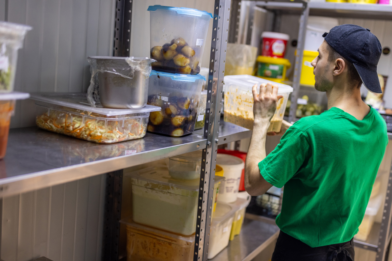 A man in a green t-shirt and a dark backwards baseball cap puts a container of food on a set of shelves with other containers of food.