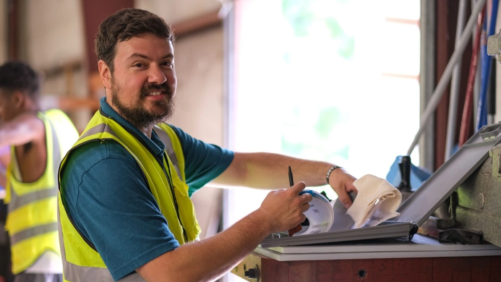 A man in a yellow safety vest works on a laptop in a workshop, representing The Industrial Commons' innovative training efforts.