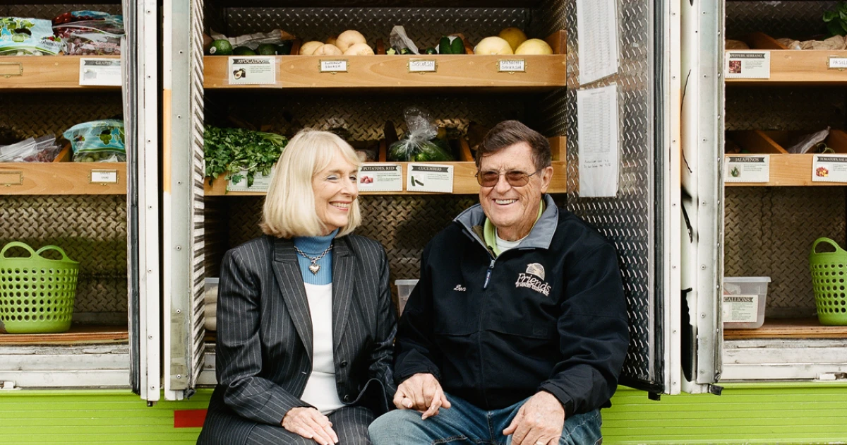 Don and Nancy Bergman hold hands and smile in front of an open door of their mobile market, which displays various fruits and vegetables.